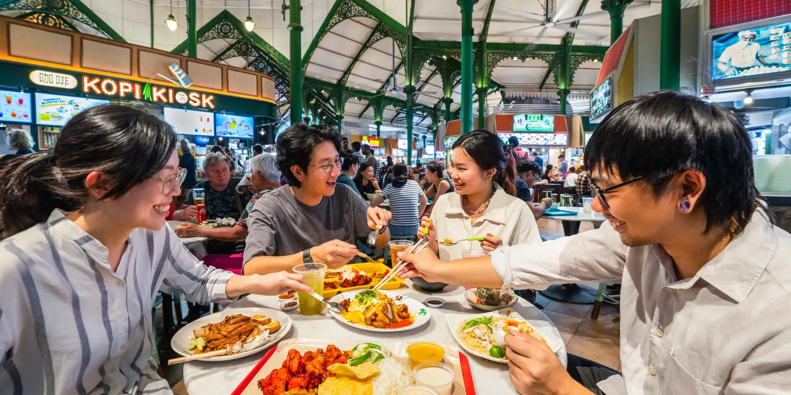 Friends sharing plates of hawker food at a table under the Victorian ironwork of Lau Pa Sat food centre, Singapore