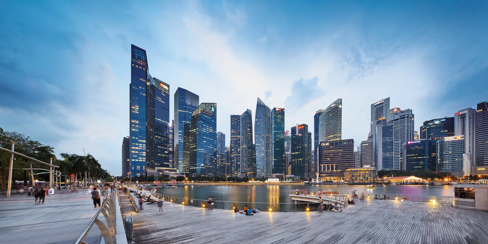 Singapore's CBD skyline at dusk seen from the free Marina Bay waterfront promenade, with people walking and city lights reflecting on the water