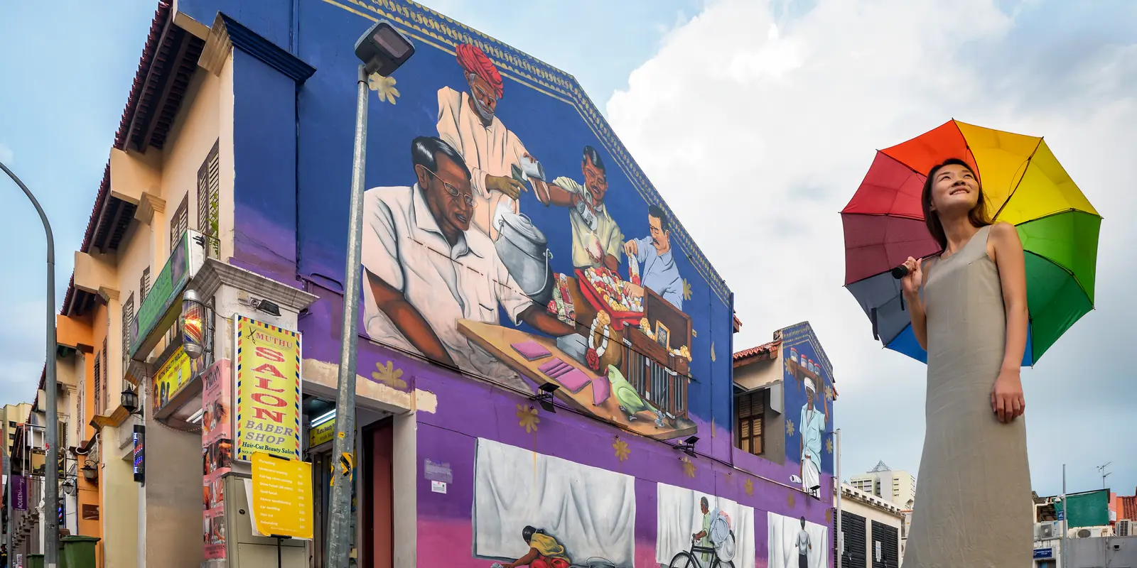 A woman holding a rainbow umbrella in front of the Traditional Trades of Little India wall mural, under a partly cloudy Singapore sky